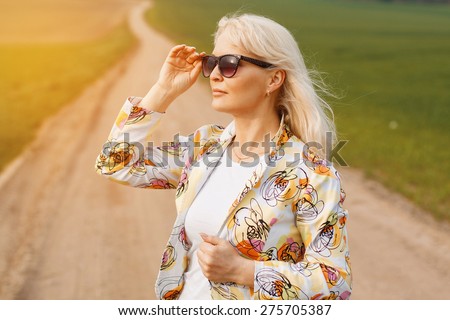 Aged woman in a jacket on the road in the green field. Holds sunglasses.