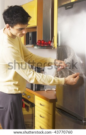 Caucasian dark-haired man in yellow sweater taking cellophane off from the refrigerator in the kitchen