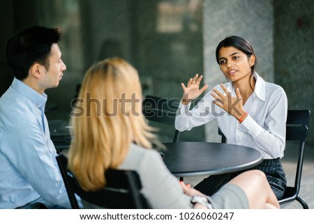 A young and attractive Asian Indian woman is interviewing for a job. Her interviewers are diverse -- one is a Chinese man, the other a Caucasian woman. The Indian woman is gesturing as she talks.