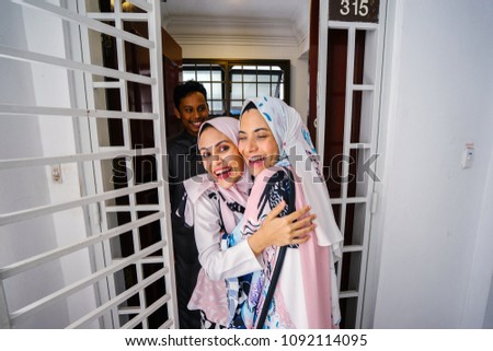 A Muslim husband and wife couple greet a guest at their door and invite her in to celebrate Hari Raya, a Muslim celebration of love and forgiveness. The two women are hugging joyously as they meet.