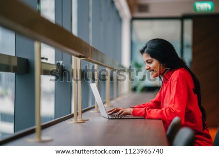 A young and attractive Asian Indian student woman works on her notebook laptop at a wooden desk during the day. She is focused and typing on her notebook; the very image of productivity.