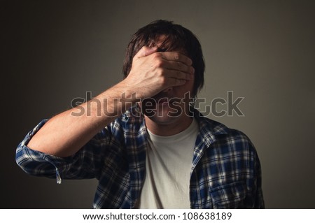 Ashamed man covering his eyes with hand, casual clothing, low light. Face palm, shame, problem, trouble, ignorance concept.