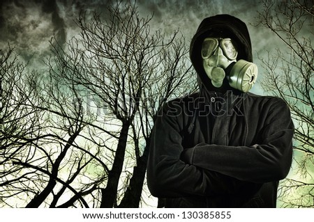 Man in dark clothes wearing a classic gas mask respirator, tree branches in the background.