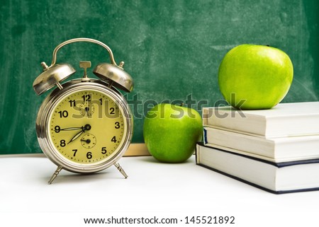 School time again. Clock, apples and books on teacher's table, green chalkboard in background. Back to school.