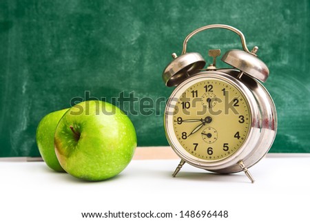 Vintage alarm Clock and green apples on teacher\'s table, green chalkboard in background.