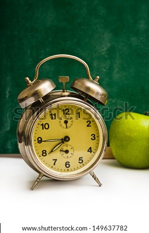 School time again. Clock and apples on teacher\'s table, green chalkboard in background. Back to school.