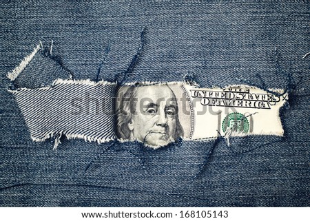 Hundred dollars bill through torn blue jeans texture. American economy concept.