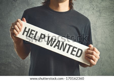 Casual man asking for help holding white board with title Help Wanted.