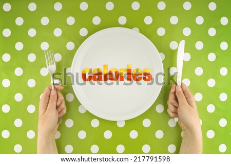 Female hands at dinner table holding fork and a knife above flat white plate with calories for a meal, conceptual dieting image.