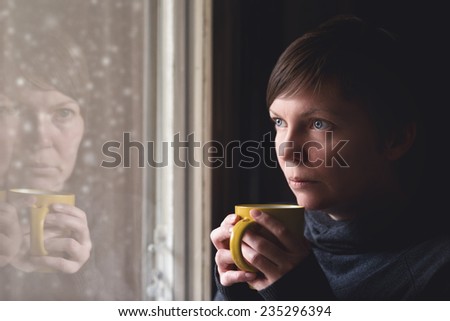 Lonesome woman drinking cup of coffee by the window of her living room, looking out at snow falling with a sad look on her face. Selective focus with shallow depth of field.