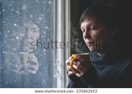 Lonesome woman drinking cup of coffee by the window of her living room while the snow is falling outside. Selective focus with shallow depth of field.