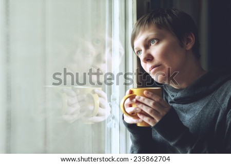 Lonely woman drinking cup of coffee by the window of her living room, looking out with a sad look on her face. Selective focus with shallow depth of field.