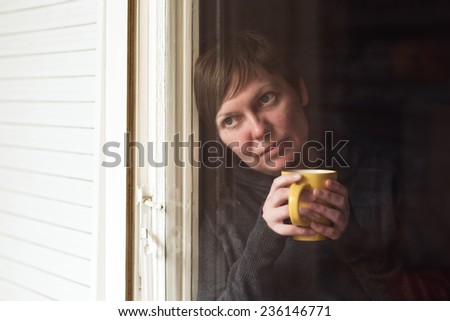 Lonely woman drinking cup of coffee by the window of her living room, looking out with a sad look on her face. Selective focus with shallow depth of field, shot through the window.
