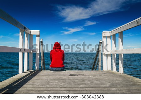 Alone Young Woman in Red Hooded Shirt Sitting at the Edge of Wooden Pier Looking at Water - Hopelessness, Solitude, Alienation Concept