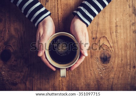 Lonely woman drinking coffee in the morning, top view of female hands holding cup of hot beverage on wooden desk, retro toned.