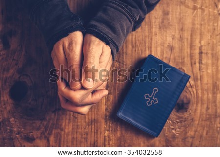 Christian man praying with hands folded and fingers crossed with Holy Bible by his side on wooden desk in church, top view