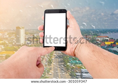 Man hand hold blank touch screen smart phone on rain drops on a window or water drops on grass blurred with cityscape background.