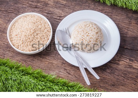 raw jasmine brown rice in white bowl and cooked jasmine brown rice on white plate with spoon and fork, on wooden table top decoration with green grass
