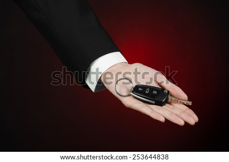 Business and gift theme: car salesman in a black suit holds the keys to a new car on a dark red background in studio