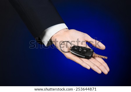 Business and gift theme: car salesman in a black suit holds the keys to a new car on a dark blue background in studio
