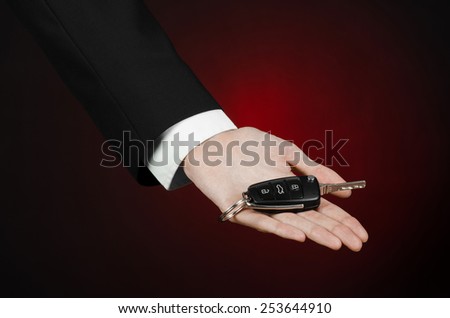 Business and gift theme: car salesman in a black suit holds the keys to a new car on a dark red background in studio
