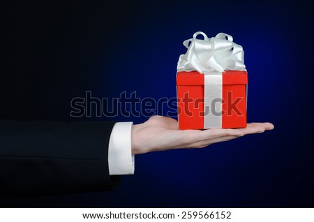 The theme of celebrations and gifts: a man in a black suit holding a exclusive gift wrapped in red box with white ribbon, beautiful and expensive gift on a dark blue background in studio isolated