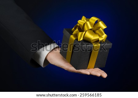 The theme of celebrations and gifts: a man in a black suit holding a exclusive gift packaged in a black box with gold ribbon, beautiful and expensive gift on a dark blue background in studio isolated