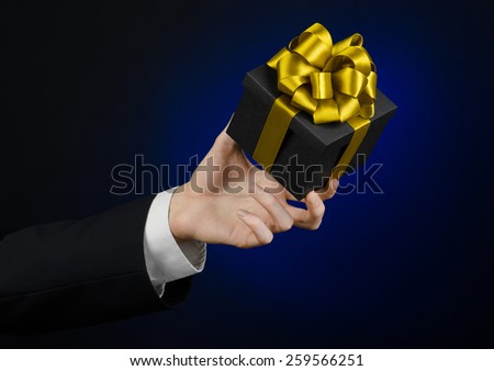 The theme of celebrations and gifts: a man in a black suit holding a exclusive gift packaged in a black box with gold ribbon, beautiful and expensive gift on a dark blue background in studio isolated