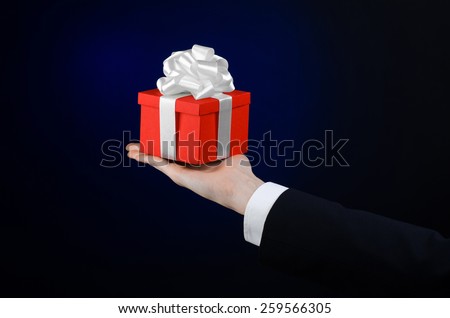The theme of celebrations and gifts: a man in a black suit holding a exclusive gift wrapped in red box with white ribbon, beautiful and expensive gift on a dark blue background in studio isolated