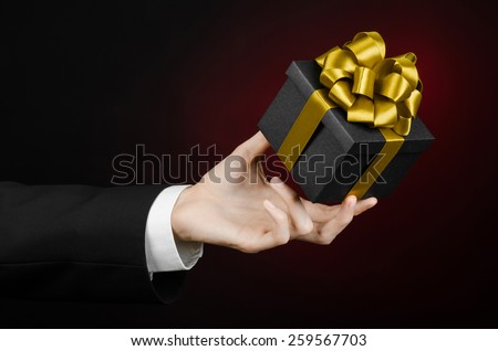 The theme of celebrations and gifts: a man in a black suit holding a exclusive gift packaged in a black box with gold ribbon, beautiful and expensive gift on a dark red background in studio isolated