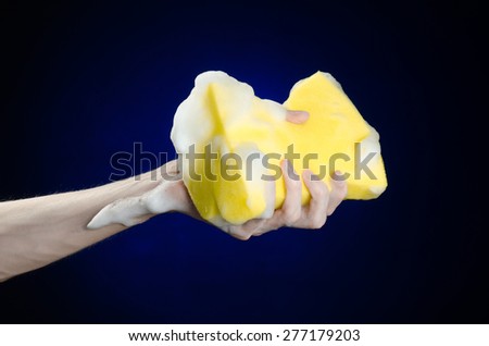 Cleaning the house and sanitation topic: Hand holding a yellow sponge wet with foam on a dark blue background in studio