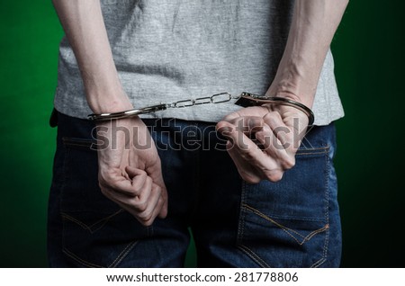 Prison and convicted topic: man with handcuffs on his hands in a gray T-shirt and blue jeans on a dark green background in the studio, put handcuffs on the drug dealer, the view from the back