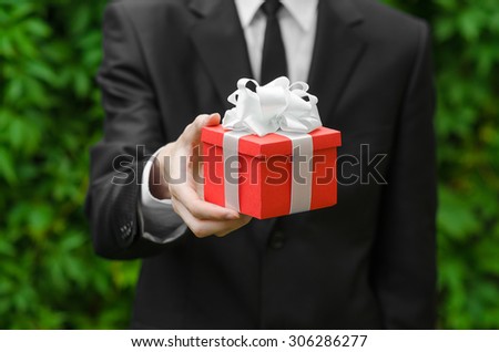 Gift and business theme: a man in a black suit holding a gift in a red box with a white ribbon on a background of green grass