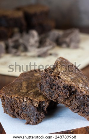 Chocolate brownies on a piece of parchment paper and chocolate chuncks on a cutting board in background.