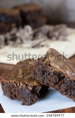 Chocolate brownies on a piece of parchment paper and chocolate chuncks on a cutting board in background.