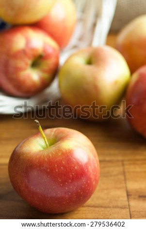 A fresh Gala apple on a wooden table with apples spilling from a white wicker basket in the background.