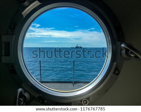 Window port of navy ship with dogs for locking the window. Outside the window also has destroyer sails beside the observer\'s ship.