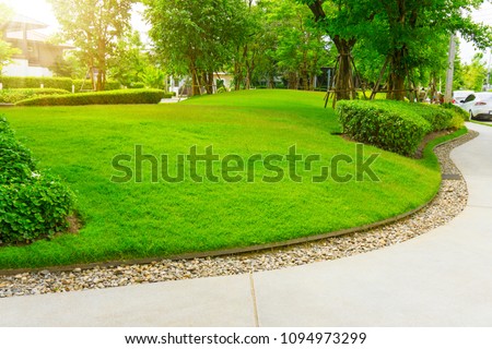 Smooth green grass lawn, trees with supporting and shrub in a good maintenance landscape and garden, gray curve pattern walkway, sand washed finishing on concrete paving, brown gravel border