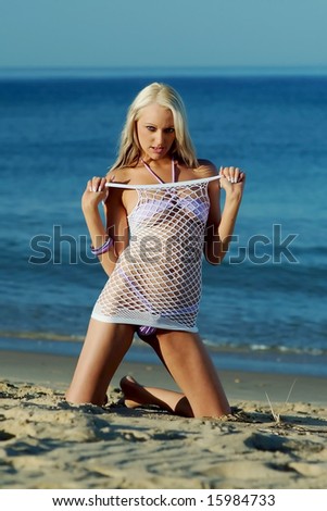 Pretty blonde girl posing on beach wearing white top & bikini.