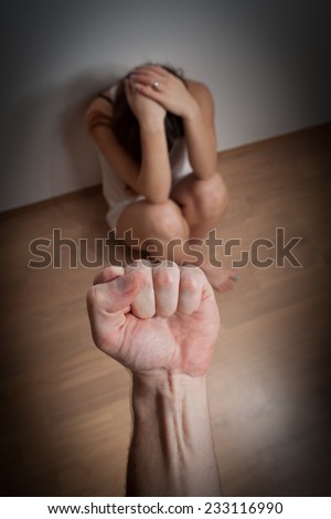 Woman/girl covering her face in fear of domestic abuse/violence sitting on the floor, punch against her face (very harsh light for underline the atmosphere, color toned image, focused on hand/punch)