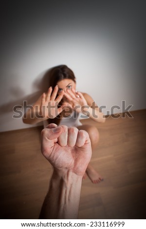 Woman/girl covering her face in fear of domestic abuse/violence sitting on the floor, punch against her face (very harsh light for underline the atmosphere, color toned image, focused on hand/punch)