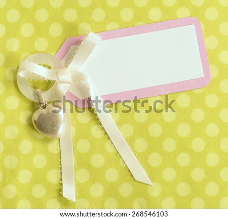 Above View of a Cute Notecard for a Baby Girl in Pink, White and Polka Dot Green Background.  A cream Satin Bow with Vintage, Silver Heart-shaped Teething Ring.  Square closeup looking down.
