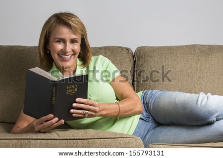 Middle-Age Christian woman comfortably sat in thew sofa reading the Holy Bible.