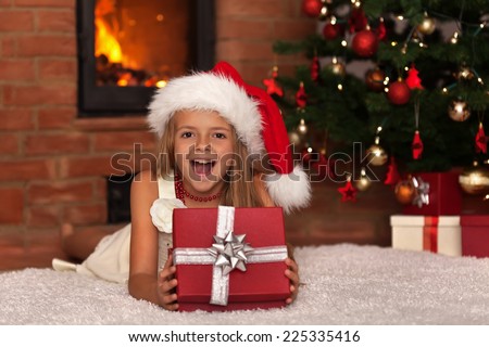 Happy little girl with Christmas present laying by the fire in front of xmas tree