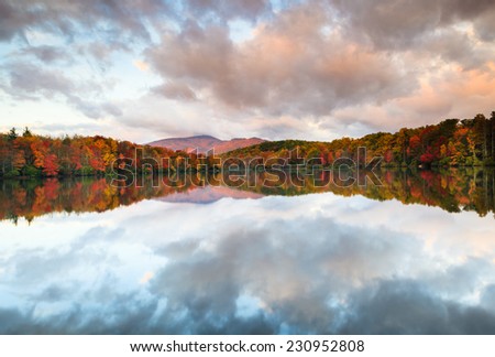 Clouds splatter across the morning sky and reflect in the water of Julian Price Lake, surrounded by trees displaying red and orange autumn colors.
