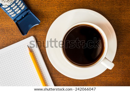 Cell phone and cup of the coffee on the wooden table with a notebook and a pencil