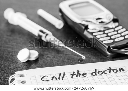 Call the doctor, medicine flows from the syringe and spread out on the table, lies next to the phone. Angle view, focus on the sign, in black and white tones