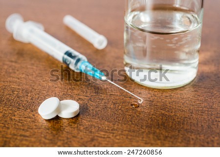 Medicine flows from the syringe and spread out on the table, lies next to a two tablets and glass of water. Angle view
