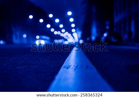 Nights lights of the big city, the night avenue with road markings and headlights of the approaching car, close up view from asphalt level. Image in the blue toning