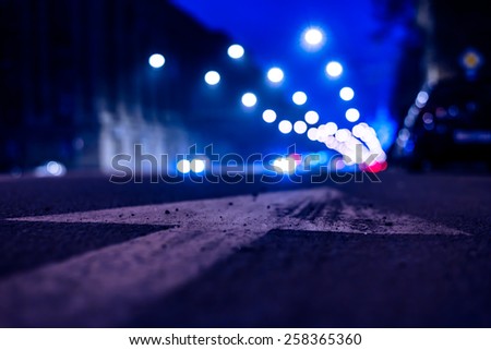 Nights lights of the big city, the night avenue with arrow and headlights of the approaching cars, close up view from asphalt level. In blue tones